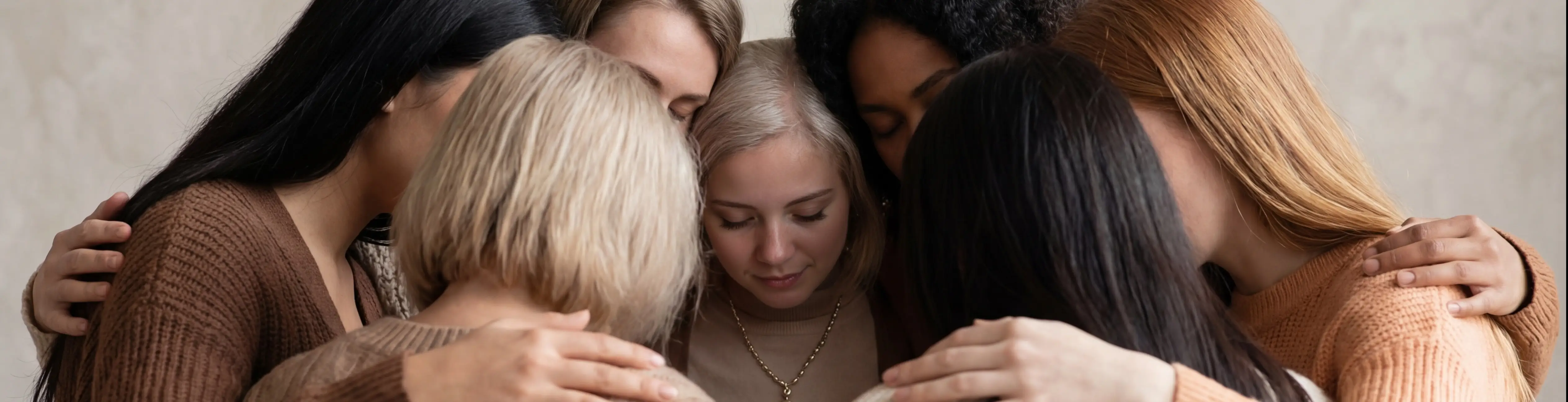 Eine Gruppe von Frauen steht eng beieinander, die Arme umeinander gelegt, die Köpfe gesenkt